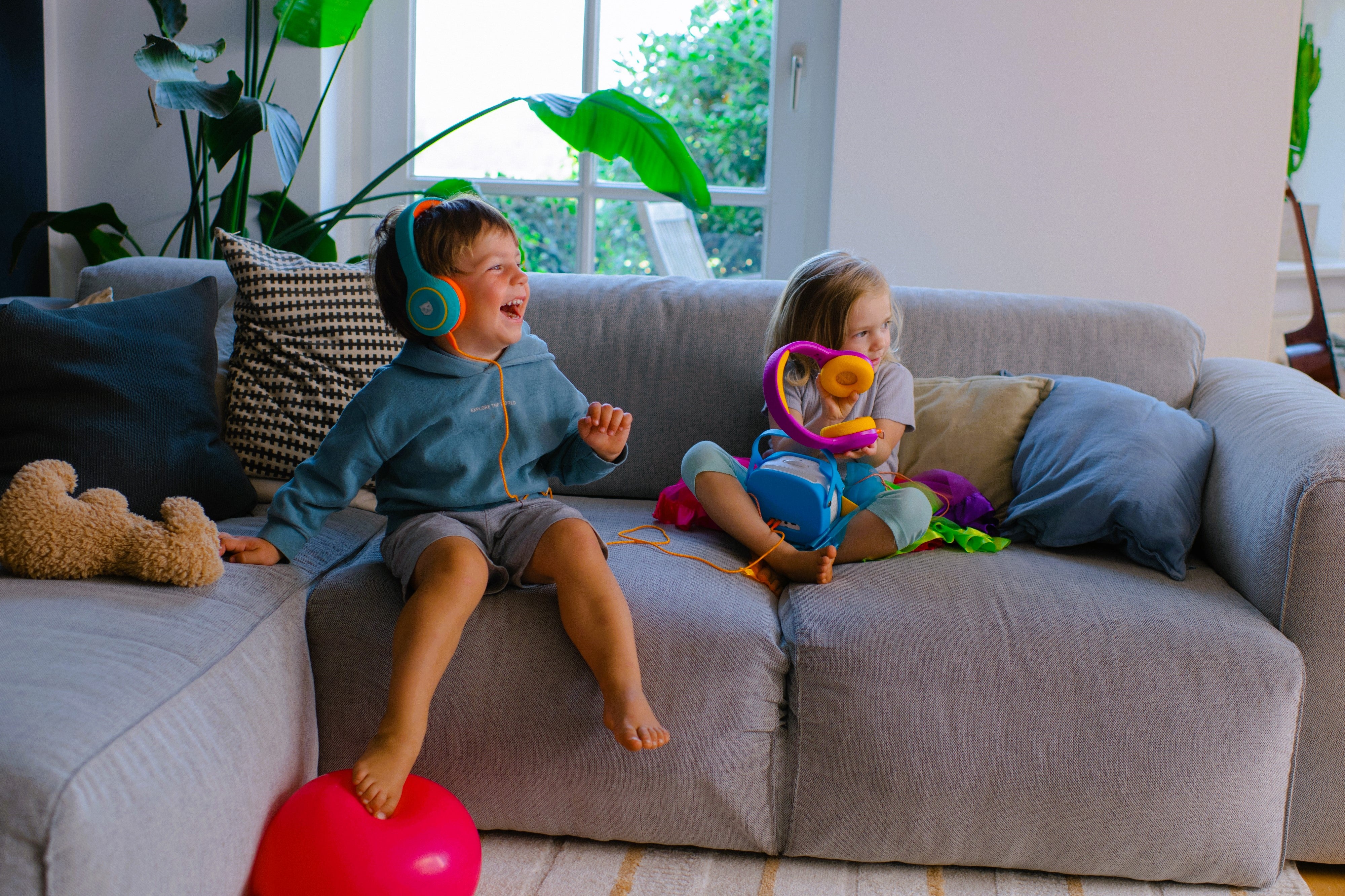 Junge und Mädchen sitzen nebeneinander auf dem Sofa und hören mit den tigerbuddies Kopfhörern etwas über die tigerbox MINI und lachen.