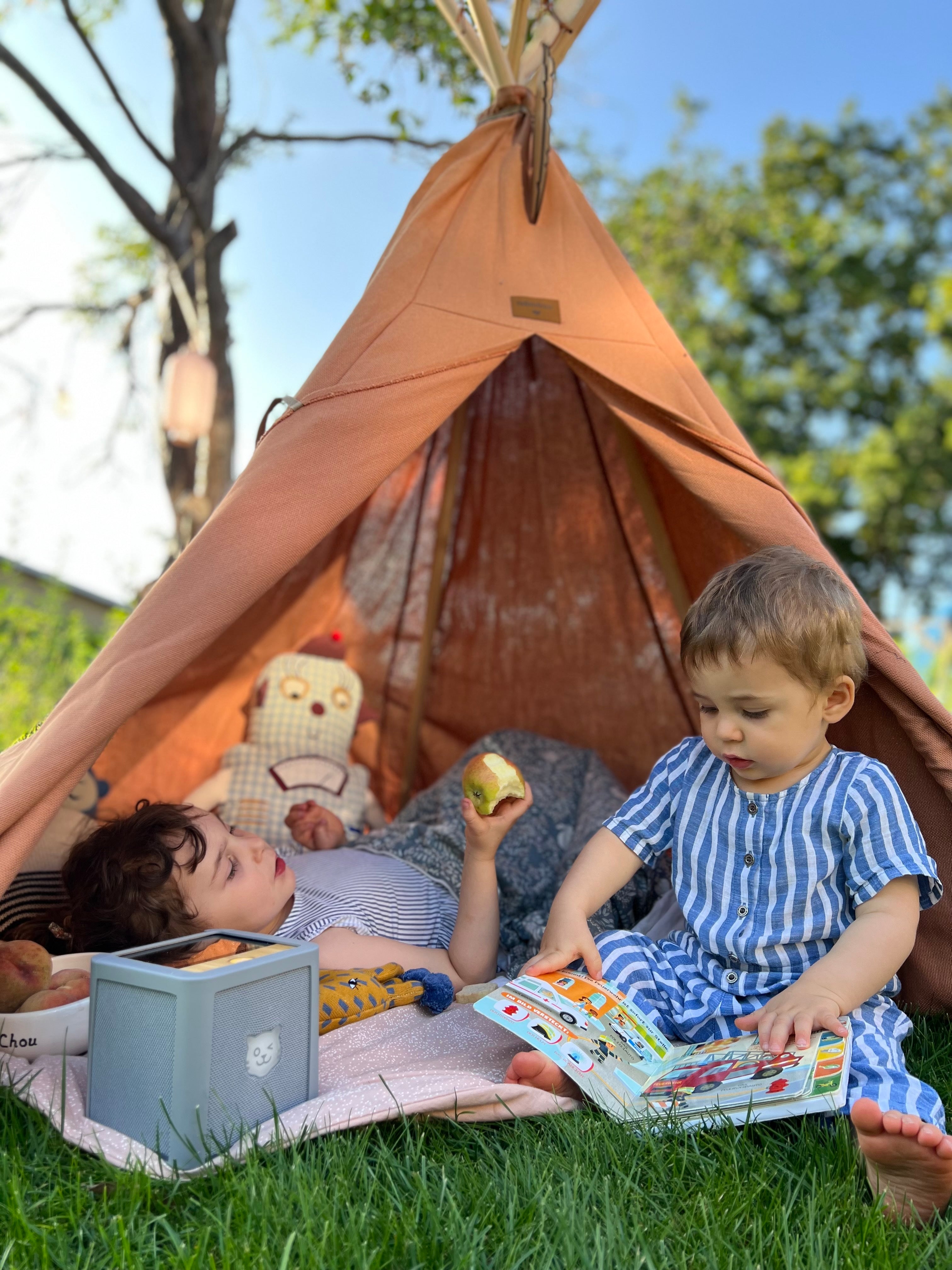 Zwei Kinder genießen ein Hörspiel auf der tigerbox TOUCH draußen im Gras und einem Tipi. Eines isst einen Apfel, das andere hält ein Buch. Decken sorgen für Gemütlichkeit.